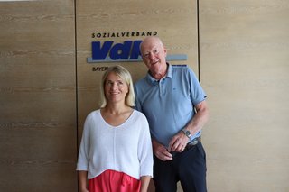 Verena Bentele und Wolf Heitland stehen nebeneinander vor einer Wand, auf der das Logo des VdK Bayern zu sehen ist. 