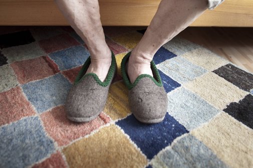 Ein alter Mann sitzt auf der Kante seines Bettes, man sieht nur seine nackten Unterschenkel, die Füße stecken in beigen Filzpantoffeln.