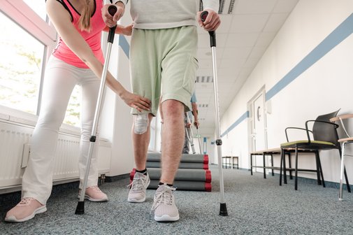 Ein junger Mann mit einer Beinverletzung w&auml;hrend der Rehabilitation, er l&auml;uft an Kr&uuml;cken, eine Frau, wahrscheinlich eine Physiotherapeutin, leitet ihn an.