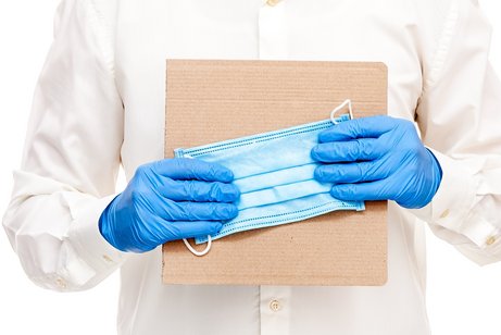 Ein Mann im wei&szlig;en Hemd tr&auml;gt blaue Einweg-Handschuhe. Vor der Brust h&auml;lt er einen schlichten Pappkarton und eine medizinische Schutzmaske.