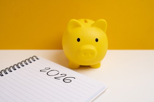 Renten 2026 Ein gelbes Sparschwein steht auf einem weißen Tisch, auf dem ein Notizbuch mit dem Eintrag "2026" liegt.