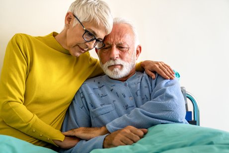 Eine Frau umarmt einen &auml;lteren Mann, der in einem Pflegebett sitzt. Beide schauen bedr&uuml;ckt.