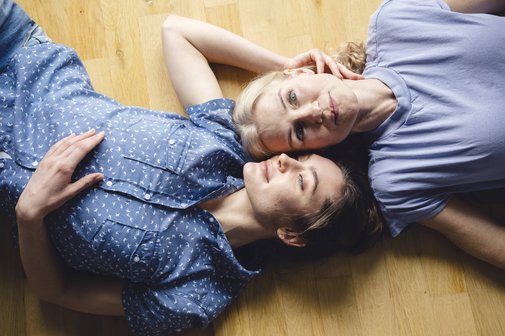 Eine Mutter und ihre erwachsene Tochter liegen gemeinsam entspannt auf dem Fußboden, sie haben die Köpfe dicht beeinander, wirken sehr vertraut und harmomisch. Die Tochter hat die Augen geschlossen, die Mutter blickt in die Kamera.