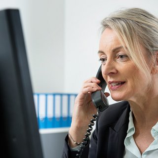 Eine Frau am Computer, sie telefoniert. Im Hintergrund sieht man Aktenordner.