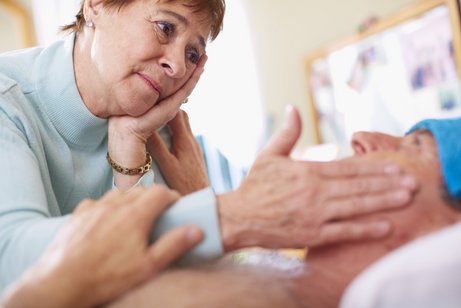 Eine ältere Frau beugt sich über ihren pflegebedürftigen Mann, legt ihm liebevoll eine Hand auf die Stirn. Sie sieht besorgt aus. 