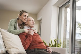 Ein Senior sitzt auf einem Sofa, schaut in die Ferne. Eine jüngere Frau, vielleicht seine Tochter, steht hinter ihm und hat liebevoll die Arme um ihn gelegt.