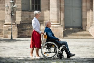 Eine Seniorin schiebt ihren Mann, der im Rollstuhl sitzt, über einen Platz