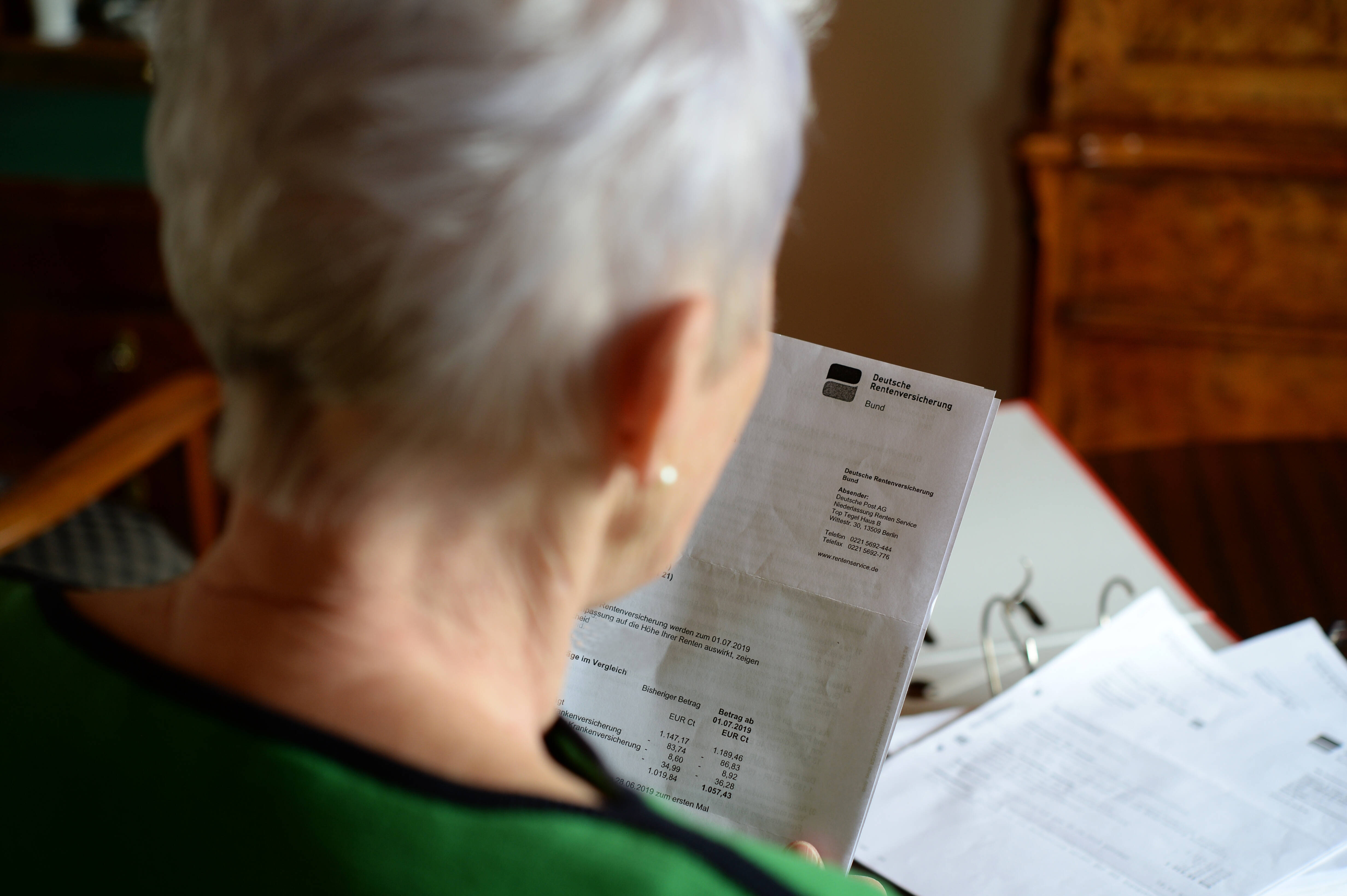 Blick über die Schulter einer älteren Frau, sie hat einen Brief der Deutschen Rentenversicherung und einen Ordner mit Unterlagen vor sich liegen.