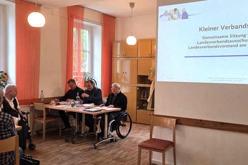 Blick in einen Raum, drei M&auml;nner sitzen nebeneinander in einem Pr&auml;sidium