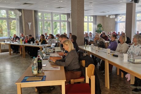 in einem Saal sitzen Frauen und Männer an Tischen und hören einem Referenten zu