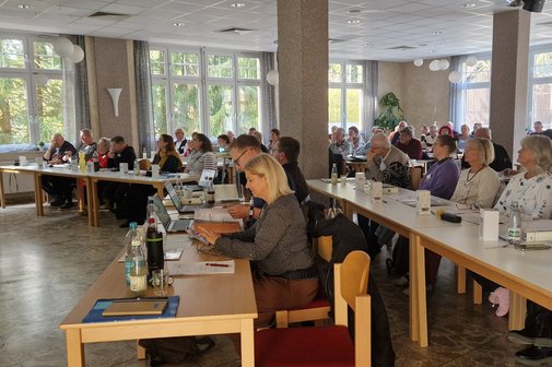in einem Saal sitzen Frauen und Männer an Tischen und hören einem Referenten zu