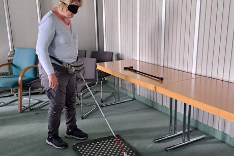eine Frau testet mit einem Blindenlangstock und einer Augenbrille ein mobiles Blindenleitsystem
