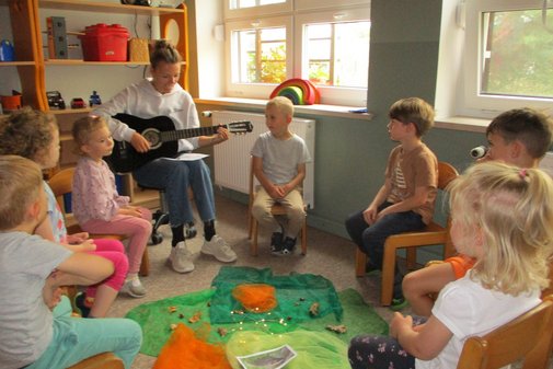 eine Erzieherin spielt Gitarre, darum sitzen Kinder im Kreis und singen