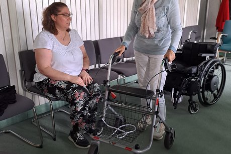 Zwei Frauen sind im Bild zu sehen, davon sitzt eine auf einem Stuhl und die zweite steht an einem Rollator.