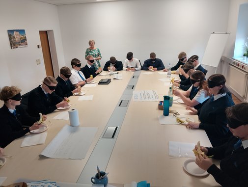 viele junge Menschen um einen Tisch mit Augenbinden, versuchen eine Banane zu essen