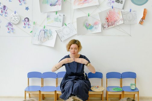 drei Kinder sitzen auf St&uuml;hlen, gegen&uuml;ber eine Lehrerin