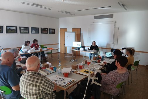 in einem Raum sitzen viele Menschen an Tischen und h&ouml;ren einer Referentin zu