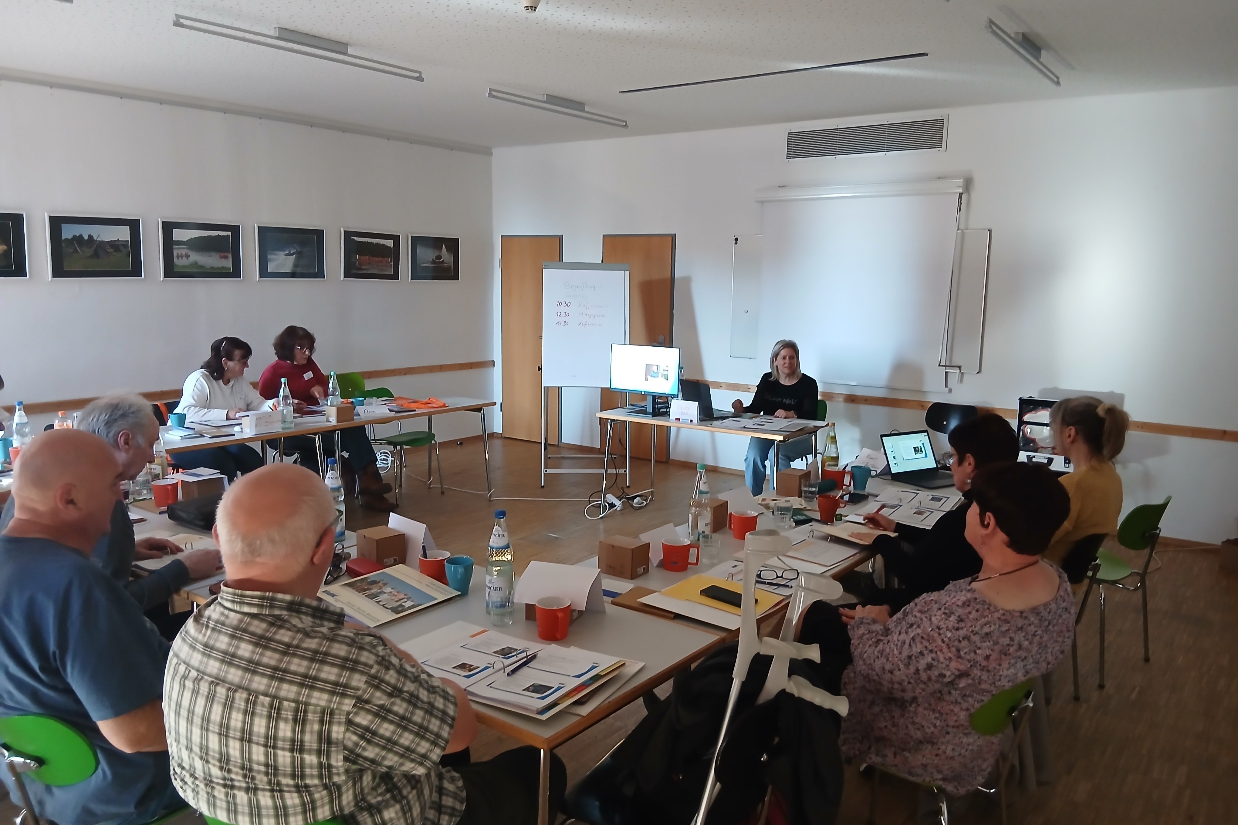 in einem Raum sitzen viele Menschen an Tischen und h&ouml;ren einer Referentin zu