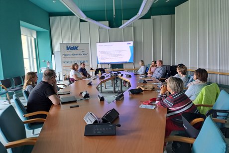 Personen sitzen in einem modernen Konferenzraum um einen großen ovalen Tisch und verfolgen eine Präsentation.