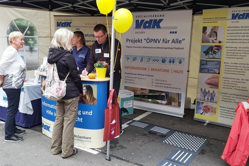 ein Infostand, der von einer Frau und einem Mann betreut wird, mit Besuchern davor