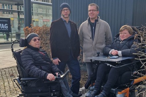 Zwei Rollstuhlfahrer links und recht und in der Mitte zwei Personen stehend