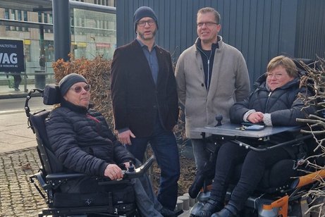 Zwei Rollstuhlfahrer links und recht und in der Mitte zwei Personen stehend