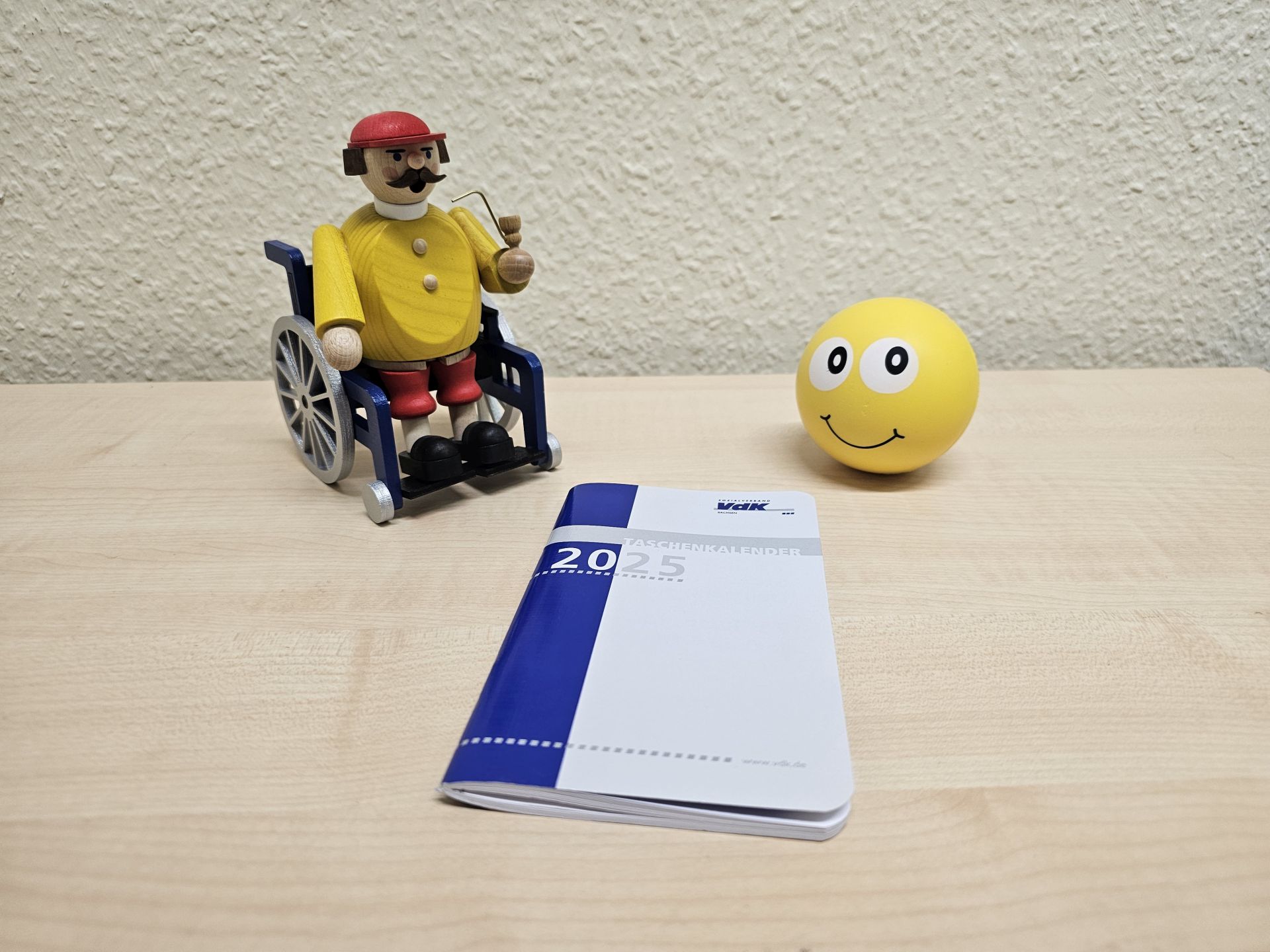 Räuchermann im Rollstuhl steht hinter einem Taschenkalender von 2025 und neben einem Ball mit Smiley